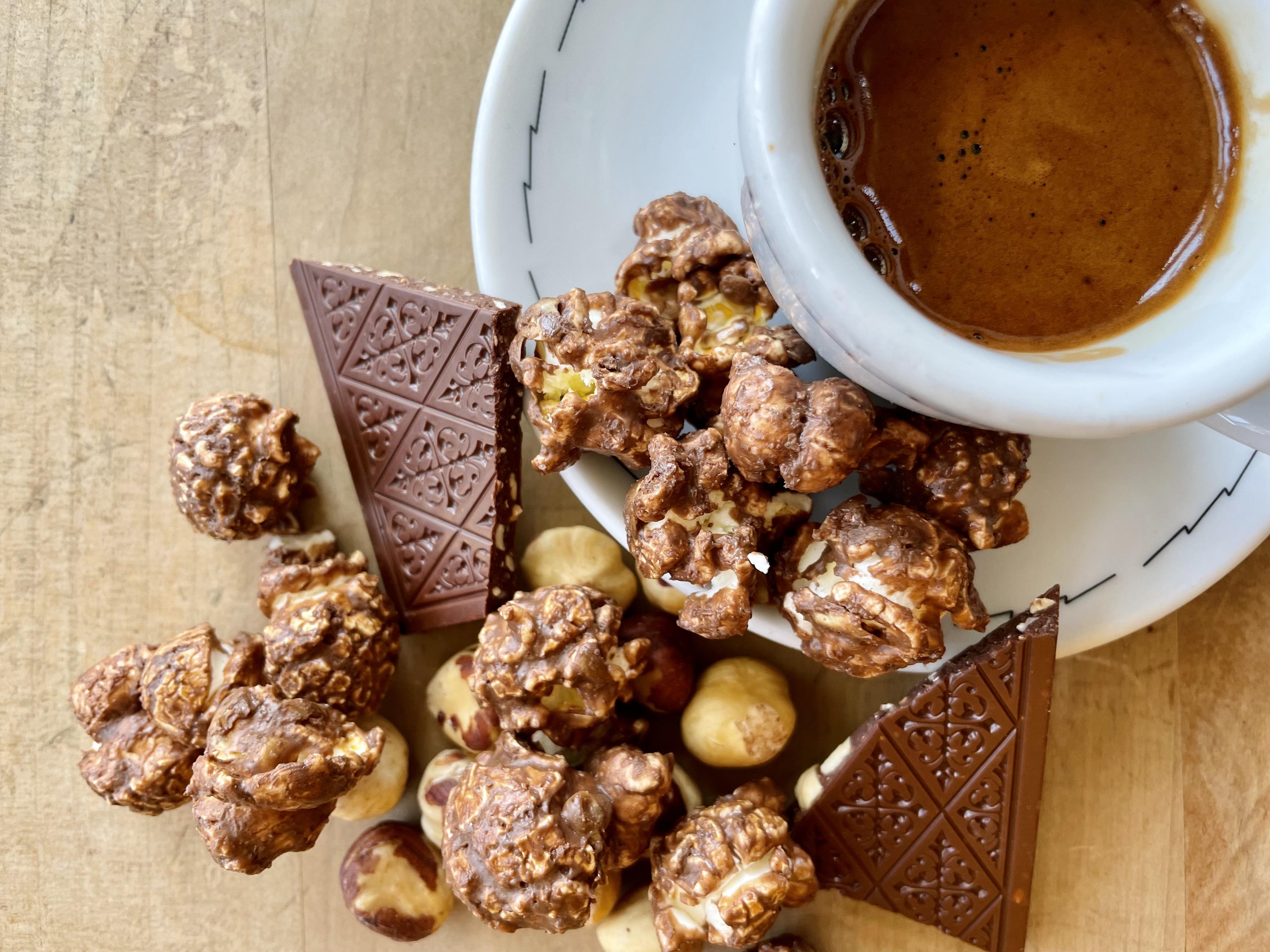 Photograph of Chocolate Hazelnut Espresso craft popcorn on a wooden table alongside hazelnuts, chocolate bar chunks, and an espresso shot