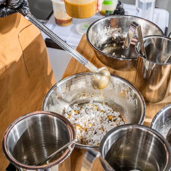 Close-up photograph of infused oil being ladled onto popcorn in a stainles steel mixing bowl.